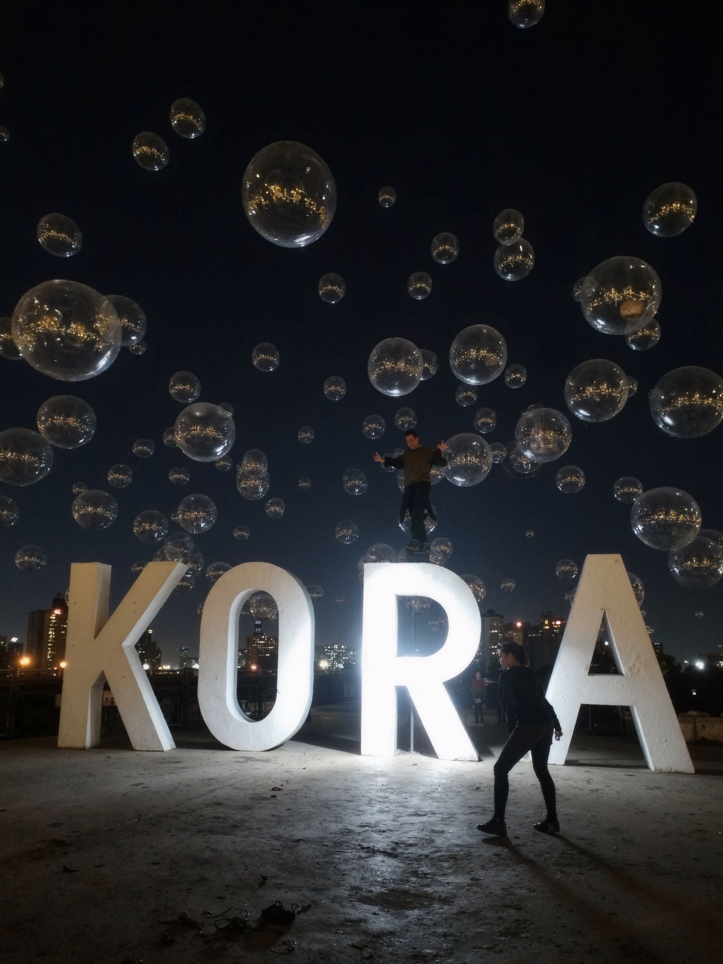 Urban back alley with a giant translucent soap bubble the size of a car, containing a little girl pushing a letter K like a swing.