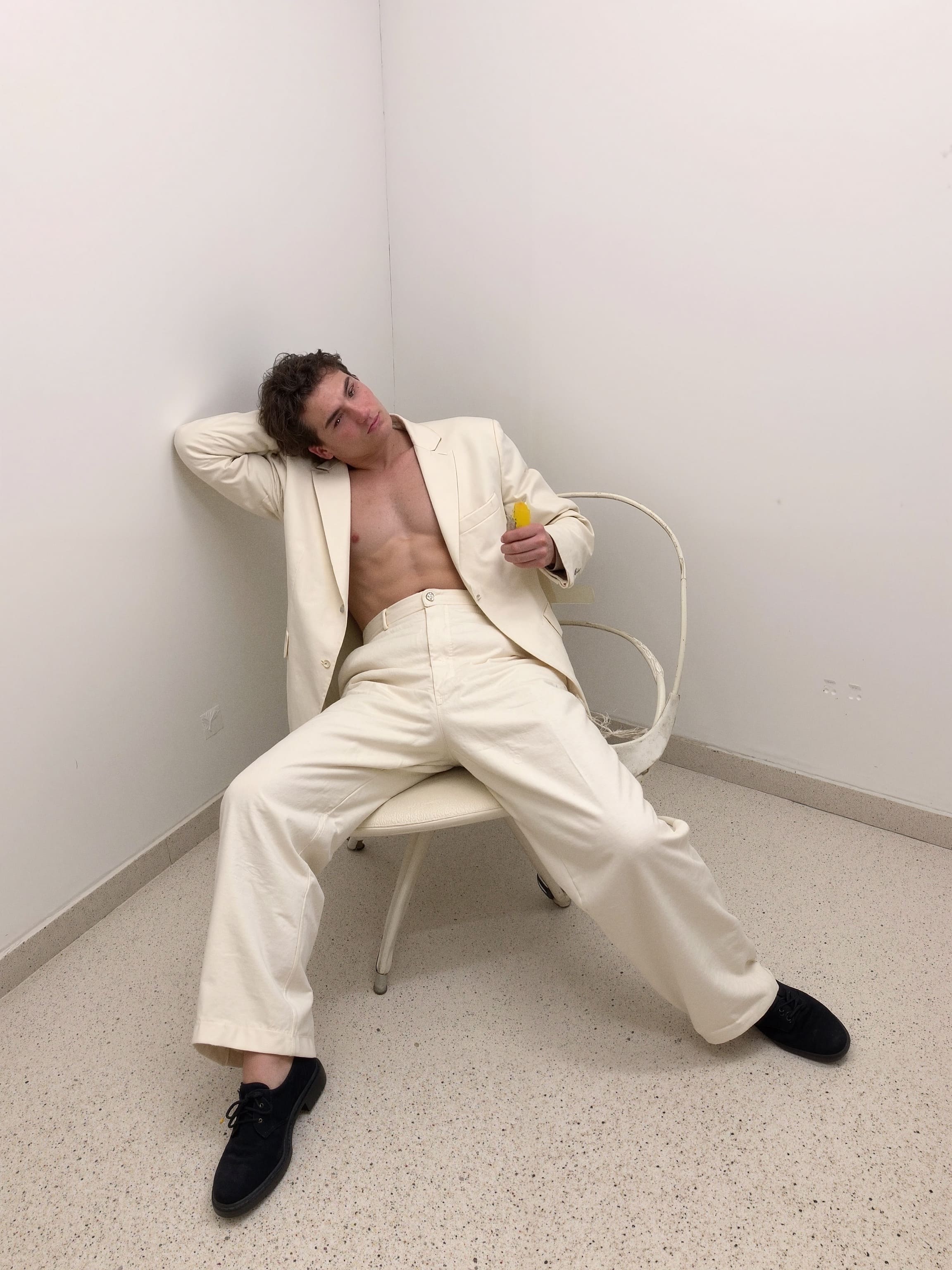 A figure slouched in a vintage Frank Gehry Wiggle Chair, wearing a loose monochrome wool suit with a half-melted popsicle.