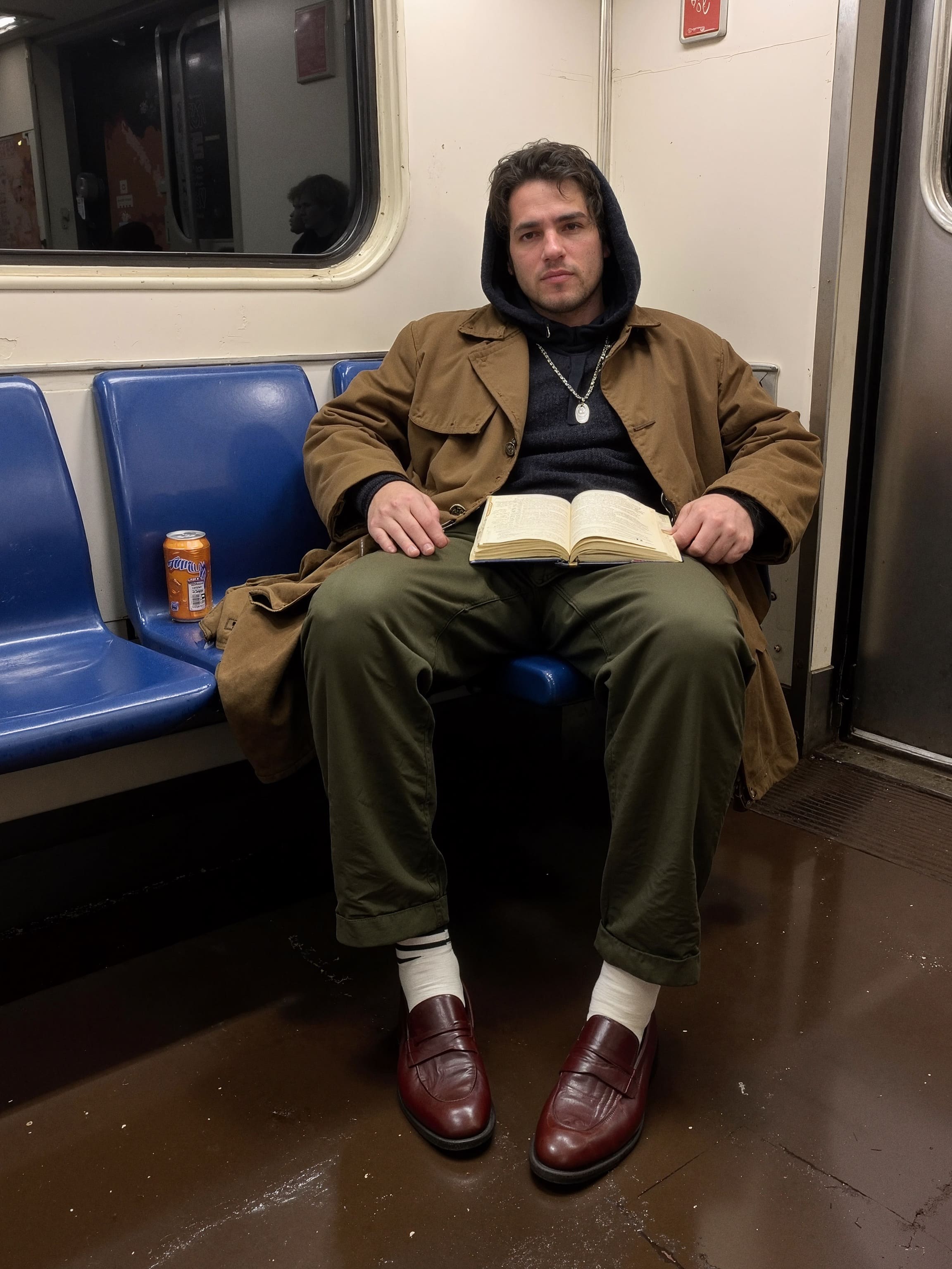 A solitary rider in a subway car wearing a heavy knit hoodie under an oversized brown trench, with an unopened can of Arizona tea.